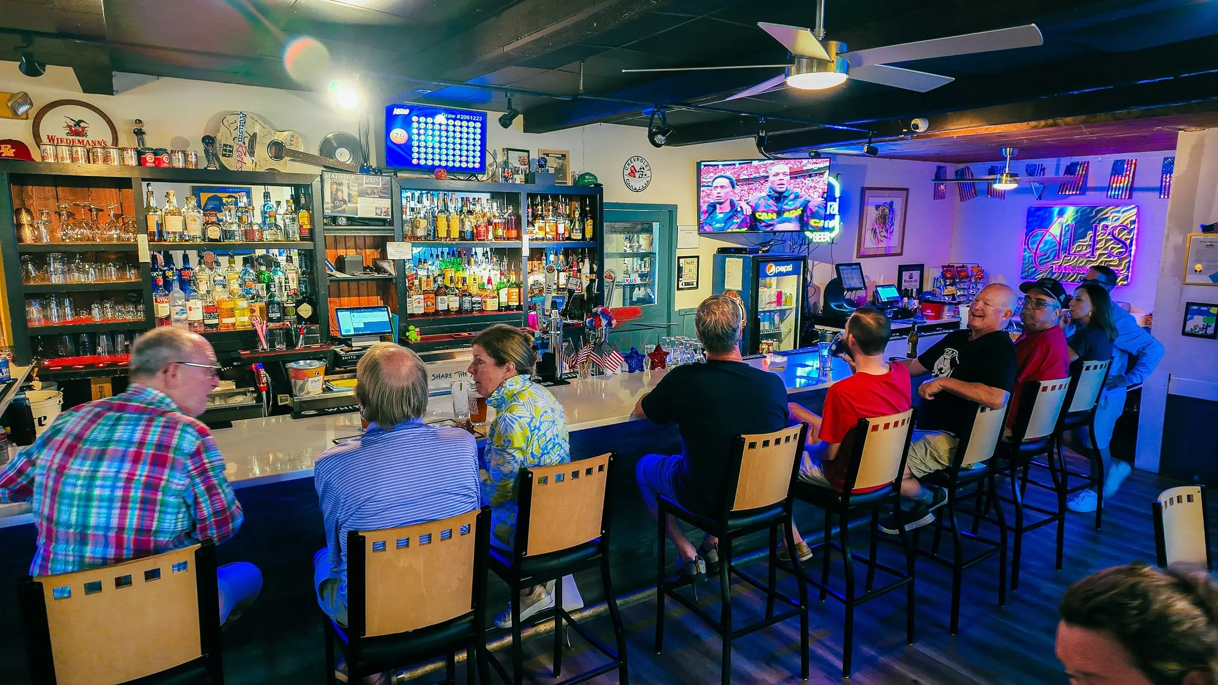 A full bar at Ali's: regulars on stools along the counter under ceiling fans, backlit liquor shelves, TVs and blue and purple neon