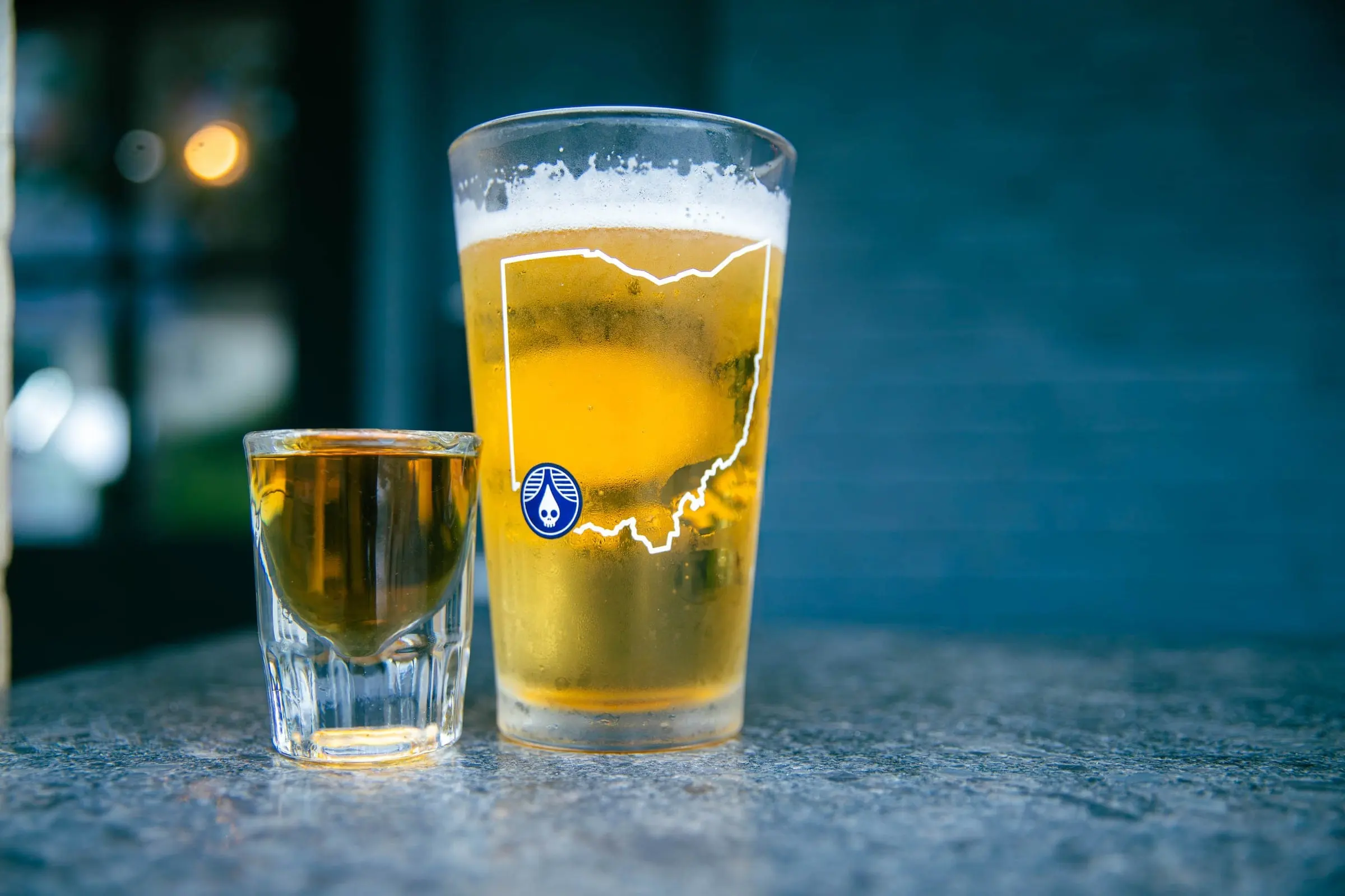 A cold pint of golden beer in an Ohio-outline glass next to a shot glass on a stone counter, against a navy background