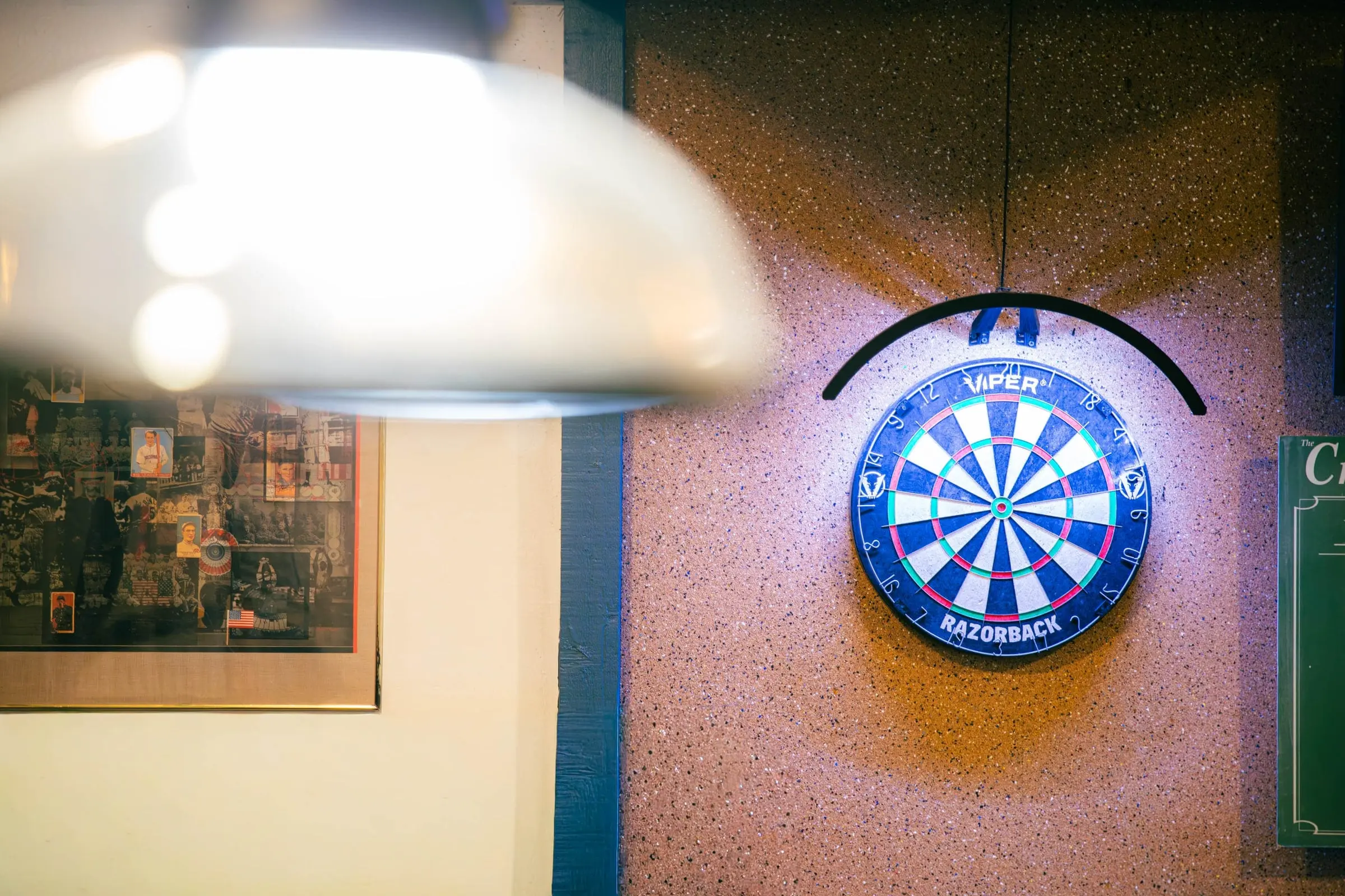 A lit electronic dartboard on the wall, framed by the soft glow of a hanging lamp