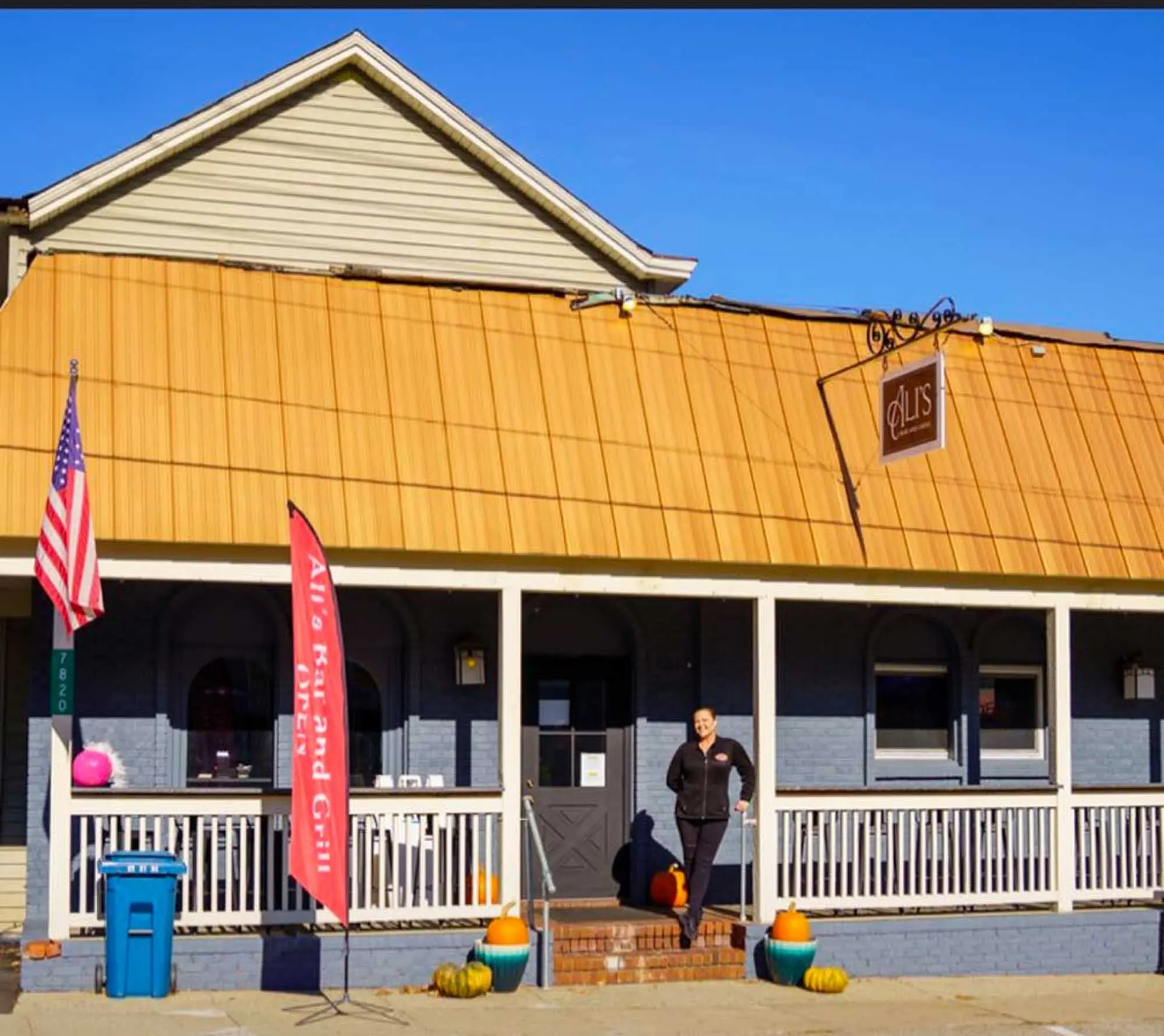 Ali's building on Camargo Road on a sunny day: copper roof, blue-gray front with a white-railed porch, an American flag, an Ali's banner and the hanging sign