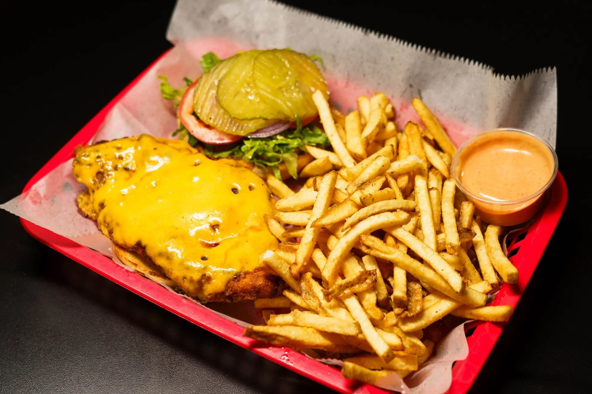 Nashville hot chicken sandwich with melted cheese and pickles in a red basket with fries and a cup of dipping sauce