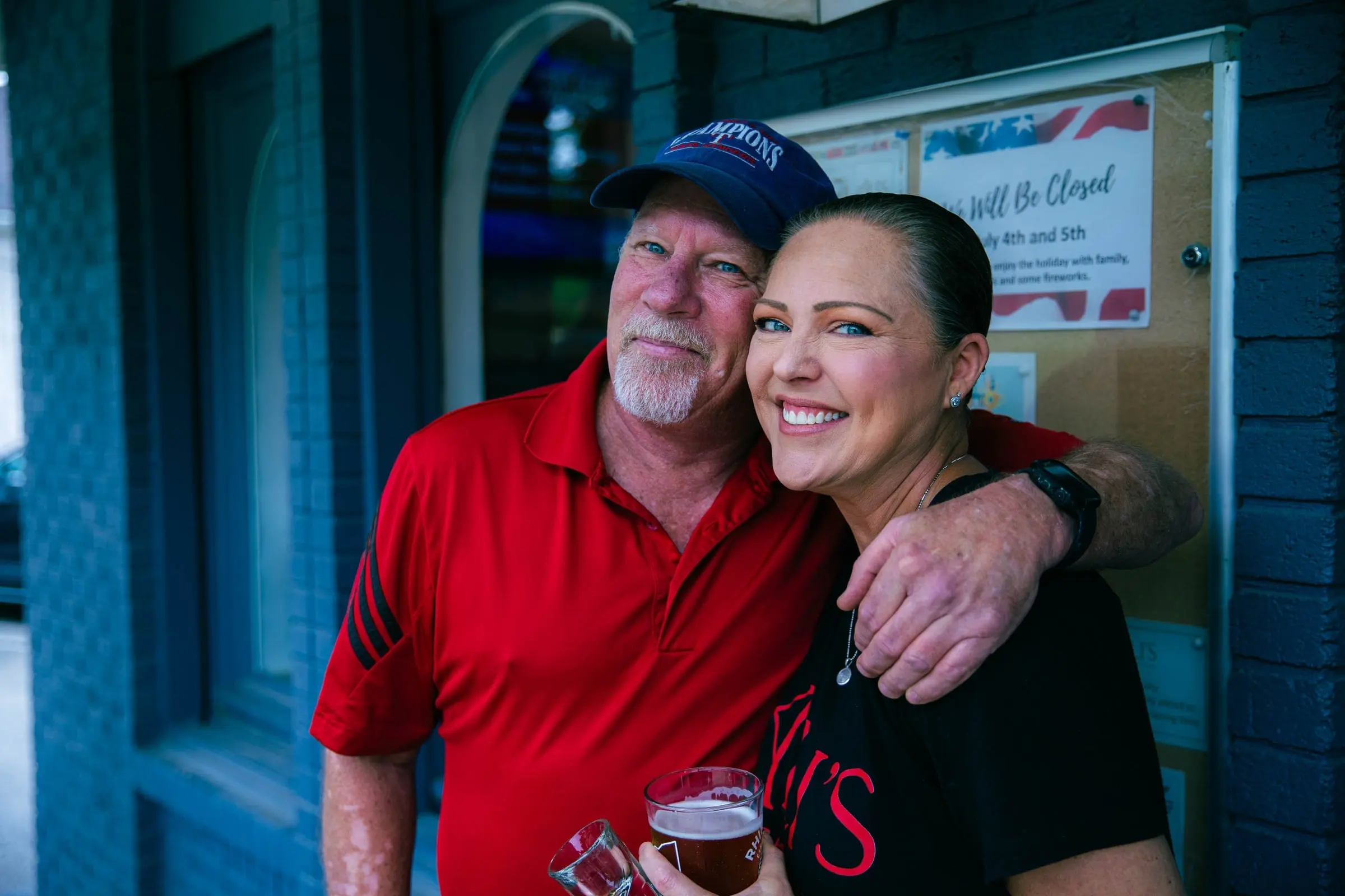 A smiling couple hug by the front door: a man in a red polo and a cap and a woman in a black Ali's T-shirt holding a beer