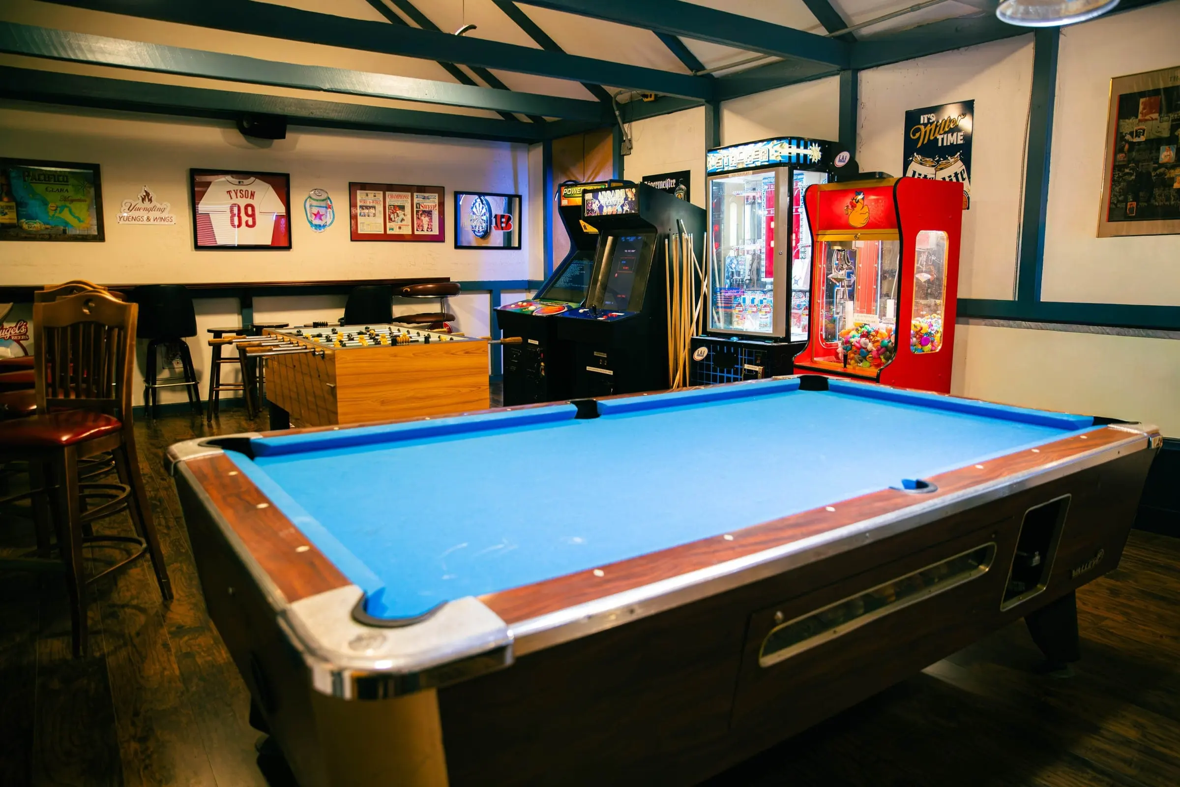 A blue-felt pool table in the game room, with foosball, arcade games and framed sports jerseys behind it