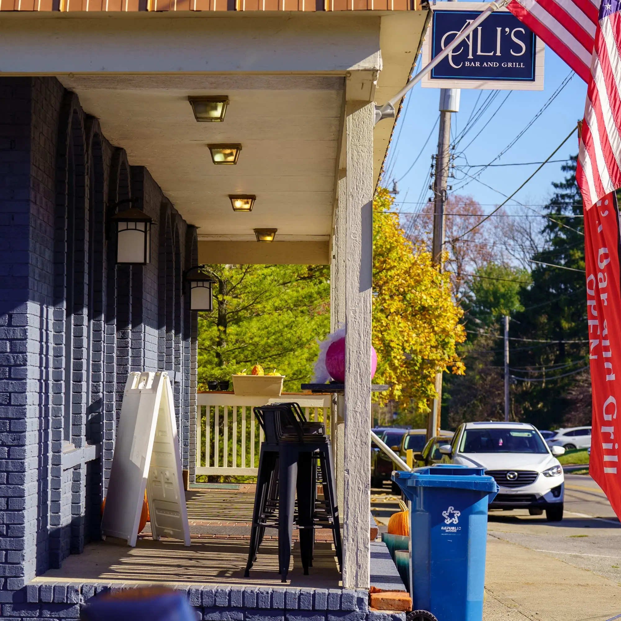The covered front porch with high-top stools, white railing, lantern lights, American flags and the Ali's sign, on a sunny fall day