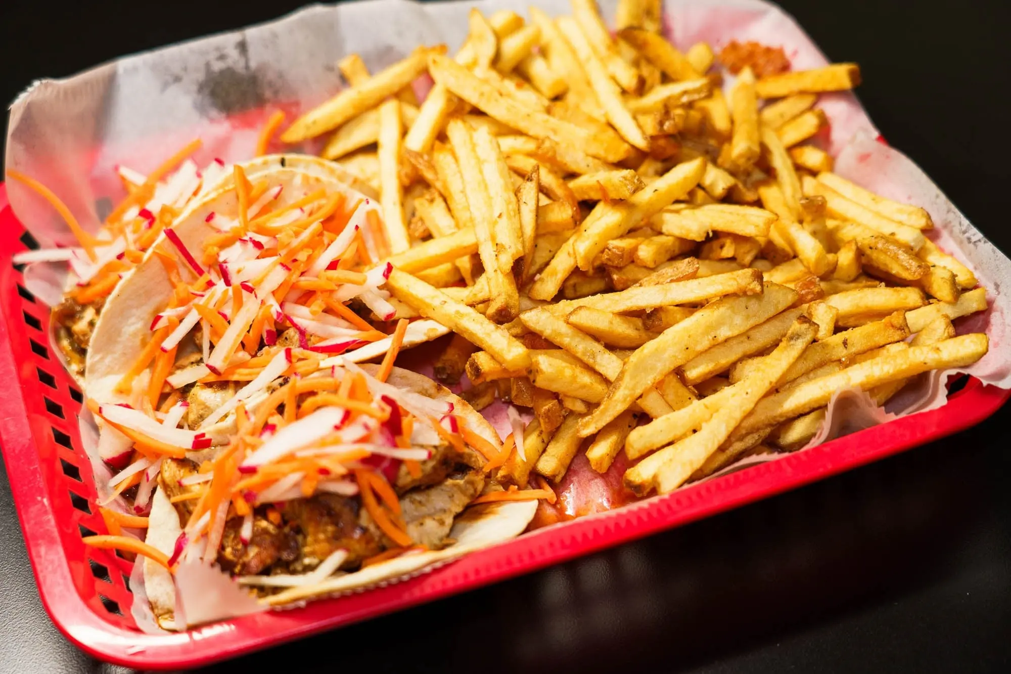 Street tacos topped with a colorful carrot and cabbage slaw beside a pile of fries in a red basket