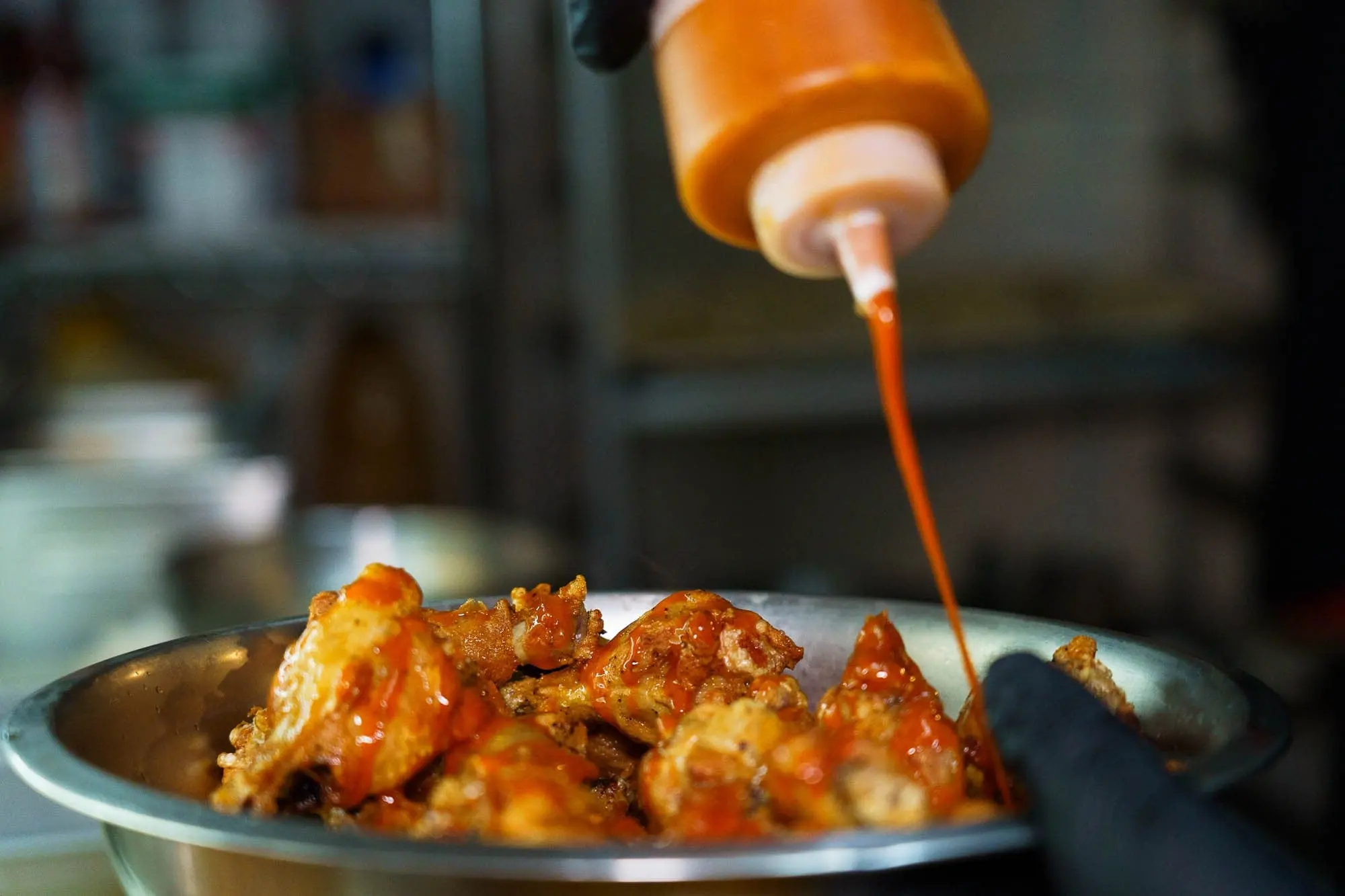 A gloved cook squeezes hot sauce from a bottle over chicken wings in a steel mixing bowl in the kitchen